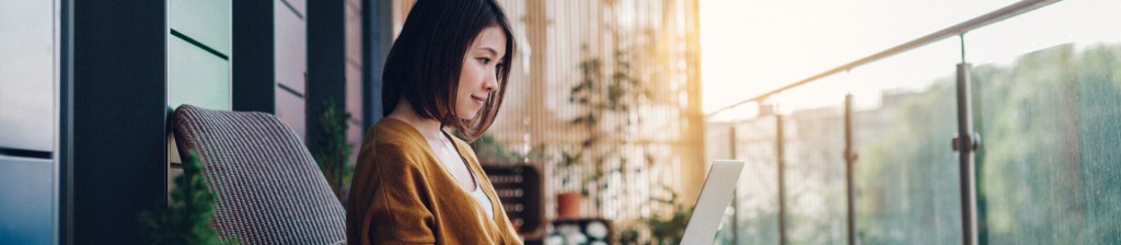 asian woman with shoulder length brunette hair sitting outside on a balcony with her laptop on her lap half smiling as she looks at her screen