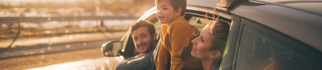 family looking out of car window, father is in driver's seat looking back at son who is perched out of the window in mom's lap, sun is hitting their face